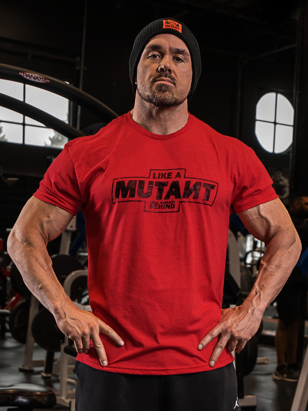 Big Ron Partlow at the gym, staring at the camera while wearing the red 'MUTANT's Truck Month' t-shirt, which features the 'Like a Mutant' phrase and 'Leave Humanity Behind' tagline in black.
