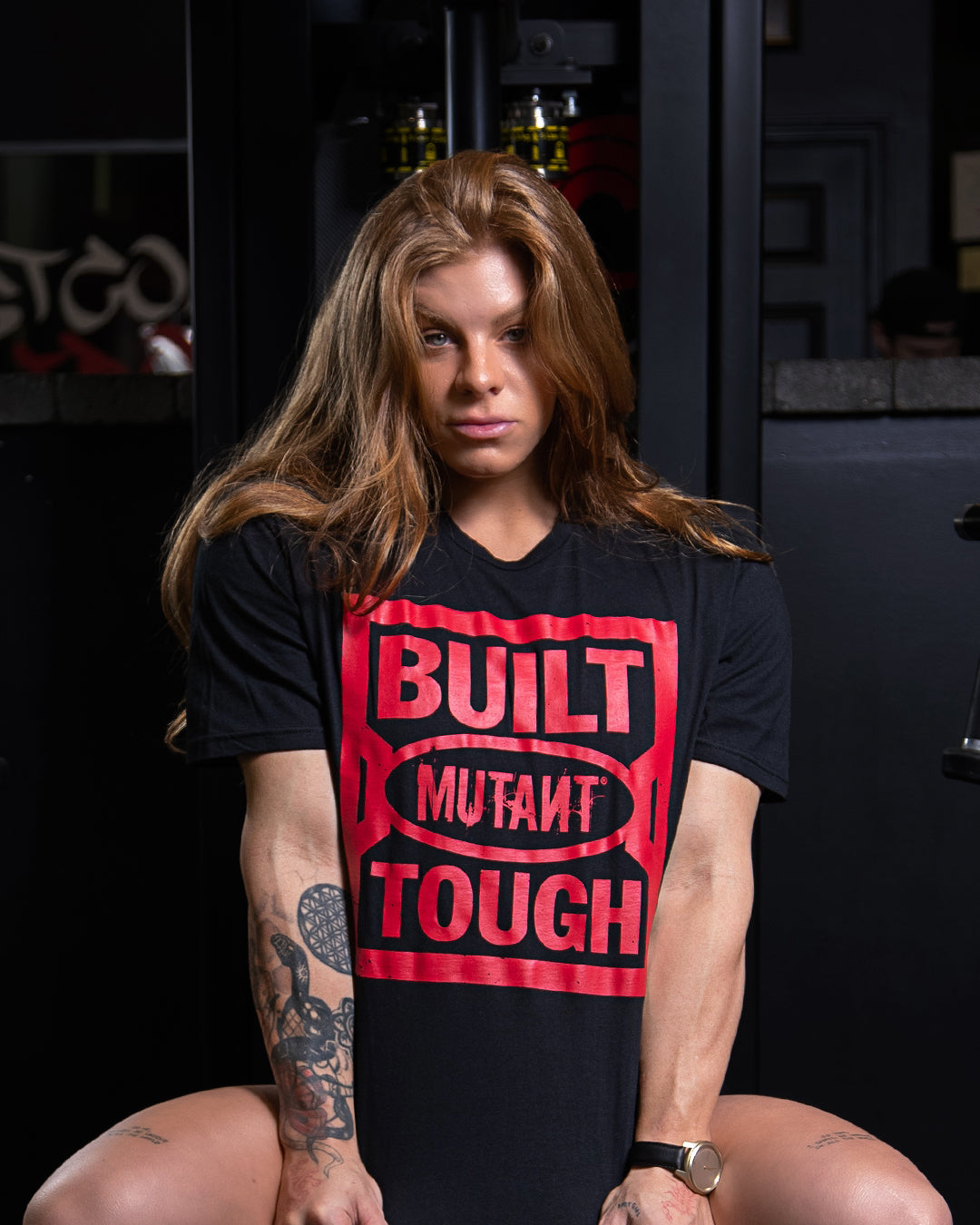 Shelby Guillaume posing at the gym while wearing the black 'MUTANT's Truck Month' t-shirt that features the phrase 'Built Mutant Tough' in red letters.