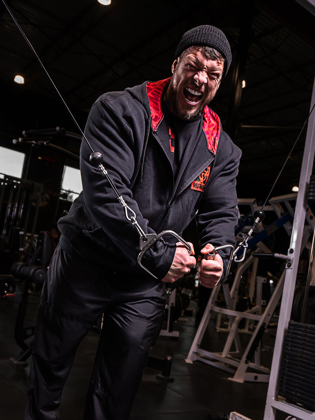Mutant athlete Jamie Christian is training in the gym wearing Black Mutant Patched Zip-Up Gym Hoodie with a red and black Mutant logo patch on left chest. Two pockets and red hood lining with red watermarked Mutant logo.