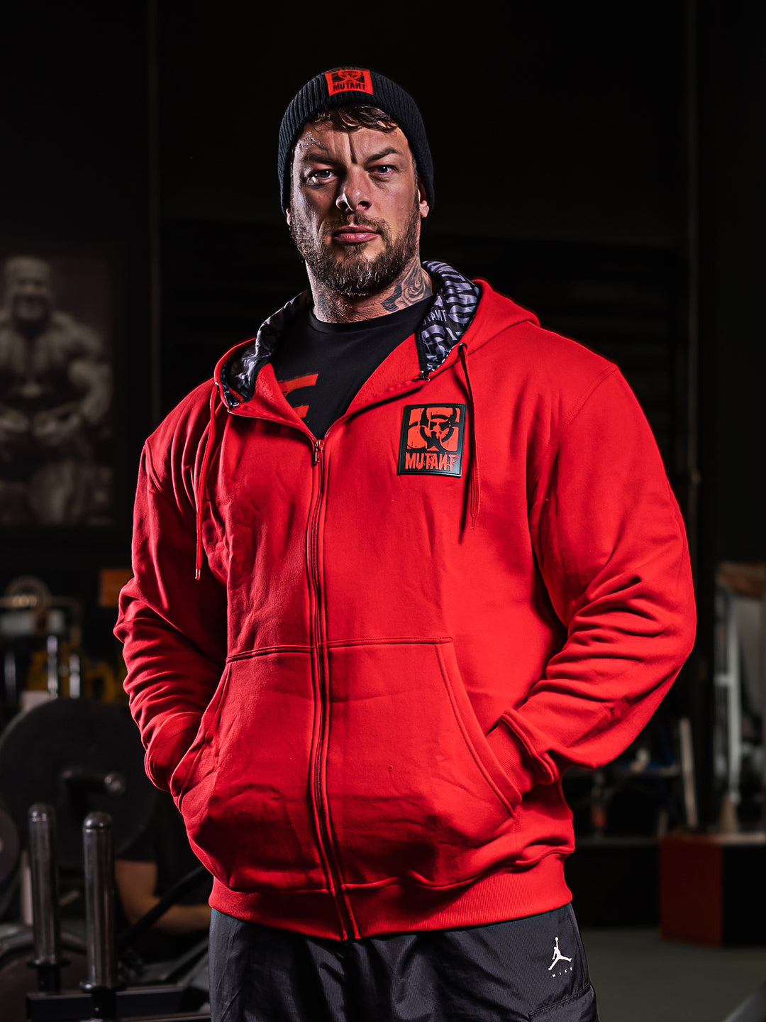 Mutant athlete Jamie Christian posing in the gym wearing Red Mutant Patched Zip-Up Gym Hoodie with black and red Mutant logo patch on left chest. Two pockets and grey hood lining with watermarked Mutant logo.