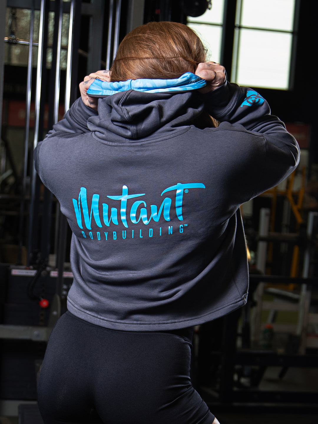Shelby Guillaume, Mutant athlete, posing in the gym to showcase the back of the livid grey Mutant Thick Script Women's Gym Crop Hoodie.