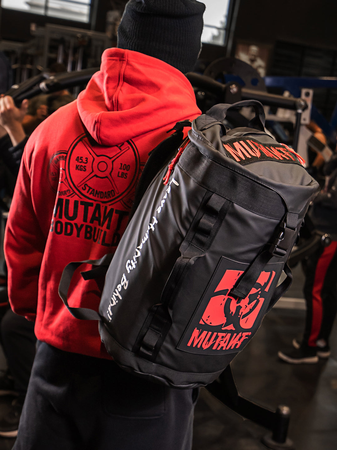 A view from behind of a man wearing a hoodie at the gym, carrying the black Military Top Load Duffel Backpack featuring a red 'MUTANT' logo.