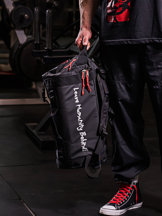 A man's hand holding the top strap of the black Military Top Load Duffel Backpack with a red 'MUTANT' logo, in a gym setting.