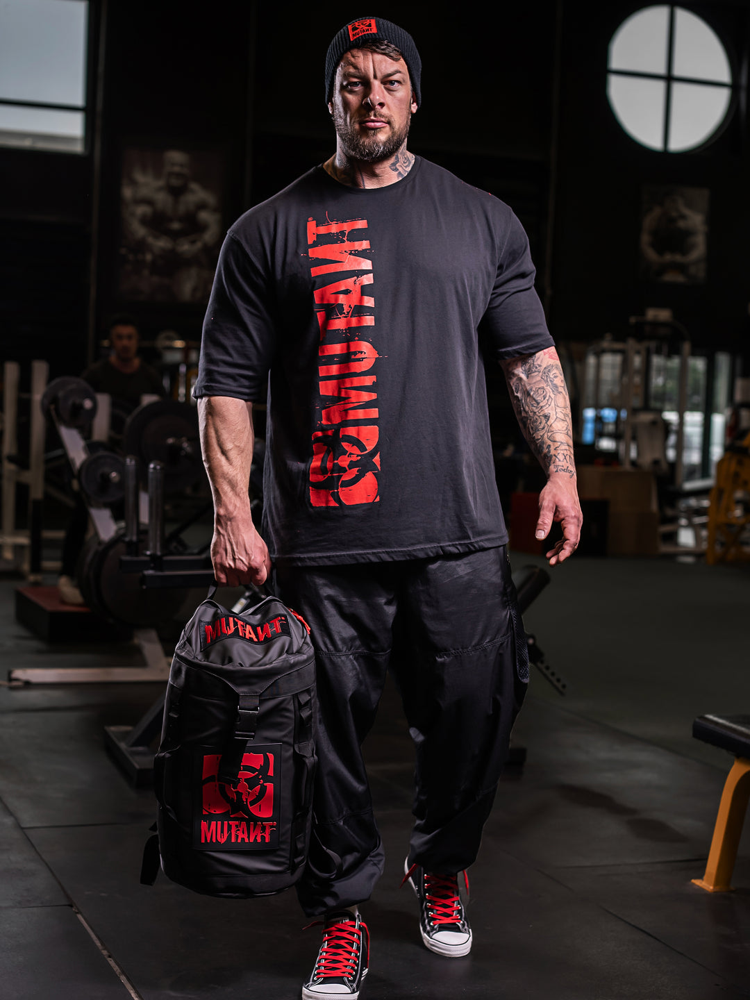Jamie Christian, the MUTANT GIANT, at the gym holding the top strap of the black Military Top Load Duffel Backpack with a red 'MUTANT' logo.